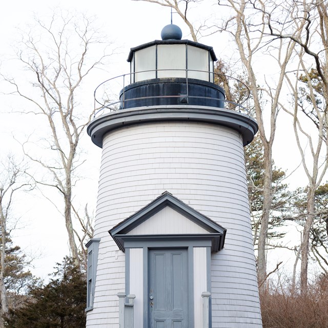 A photograph of a simple white lighthouse with wooden siding and a dormered door from the 1800s.