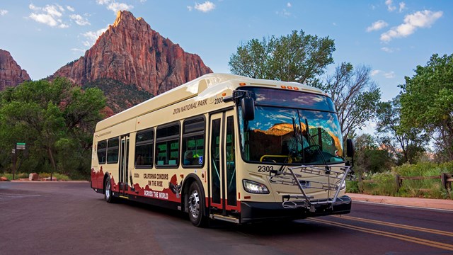 A Zion Canyon shuttle on a red road, with red sandstone formations in the background.